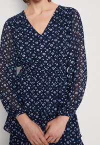 Námořnické šaty s výstřihem do V, dlouhými průsvitnými rukávy, elastickým pasem a bílými geometrickými vzory. Obsahují vrstvený volán na spodním okraji.