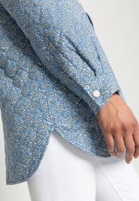 Blue floral shirt with white and green accents, long puffed sleeves, rounded hem, button closure, textured fabric. Worn with white pants.
