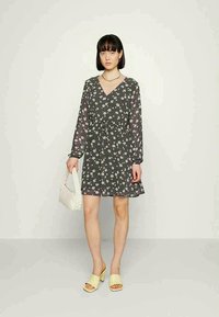 Vestido preto com flores, de mangas longas e decote em V, cintura ajustada, saia evasê; combinado com sandálias de salto amarelas e uma bolsa branca.