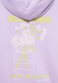 Sweatshirt lilás com um design gráfico de um frasco contendo flores. O texto inclui "A FLORECER" e "NOVA ESTAÇÃO" em amarelo. Textura suave.