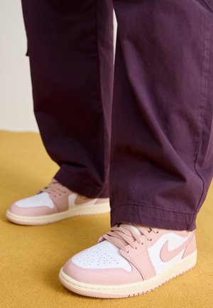 Persoon die paarse broek en roze-witte Nike sneakers draagt, staat op een gele vloer.