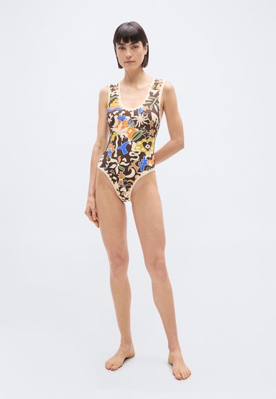 Γυναίκα που στέκεται ξυπόλητη φορώντας πολύχρωμο μαγιό ολόσωμο με αφηρημένο μοτίβο, μπροστά σε απλό φωτεινό φόντο.