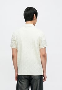 Cremefarbenes Polohemd mit strukturiertem Rautenmuster, kurzen Ärmeln und klassischem Kragen, getragen zu dunklen Jeans.