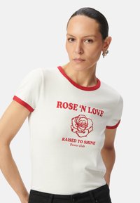 Camiseta blanca de manga corta con acentos rojos y un gráfico de una rosa con el texto "ROSE 'N LOVE" y "RAISED TO SHINE Power club".