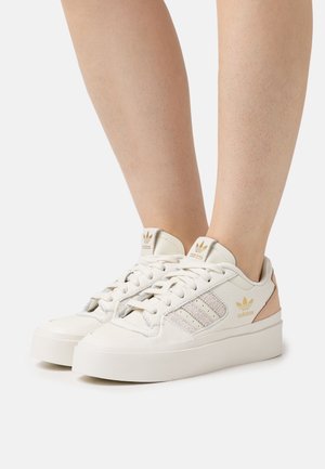 Voeten met witte Adidas sneakers met beige accenten, vastgestrikt, gefotografeerd tegen een effen witte achtergrond.