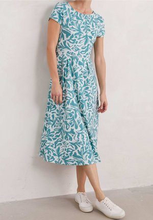 Vrouw die een teal- en witbladprint midi-jurk draagt met korte mouwen en witte sneakers, staand tegen een effen muur.