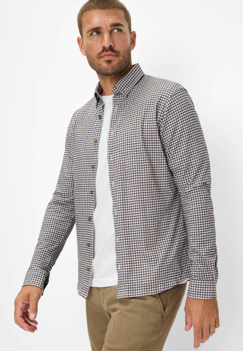 Camicia a button-up in una fantasia a quadri marrone e blu. Presenta maniche lunghe, colletto e una patta frontale. Indossata sopra una t-shirt bianca.