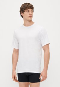 Balta medvilniniu marškinėliai trumpomis rankovėmis, apvaliu iškirpimu ir tiesiu siluetu, dėvimi su juodais šortais. Paprasta, lygi tekstūra be raštų.