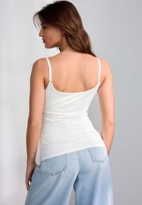 Top in tessuto bianco, aderente, con spalline sottili e scollatura a giro sul retro, abbinato a jeans a vita alta di colore azzurro chiaro.