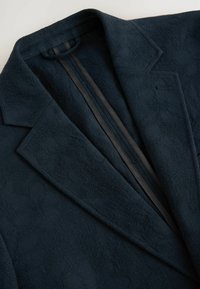 Mørkeblå struktureret blazer med et subtilt blomstermønster, der har reverskrave og sorte detaljer langs den indre søm.