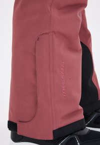Pantalons de neige rouges imperméables avec une texture lisse, présentant des accents noirs, des poignets réglables à velcro et un logo visible indiquant "VX-PRO 10000."