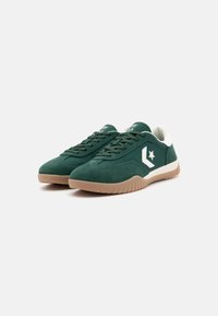 Zielone zamszowe sneakersy z białymi akcentami, z zaokrąglonym czubkiem, teksturowaną podeszwą i logo gwiazdki po boku. Sznurowadła są ciemnozielone.
