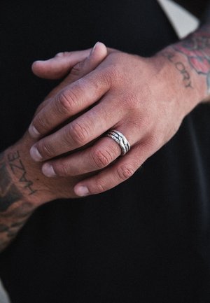 Sidabrinė suktinė juosta ant rankos su matomais tatuiruotėmis. Oda turi tekstūrinę išvaizdą, kontrastuoja su sklandžiu žiedo paviršiumi.