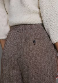 Pruunid herringbone-stiilis püksid tekstuuriga viimistlusega, nähtava nupukinnitusega ja voltidega detailidega vöökohtadel.
