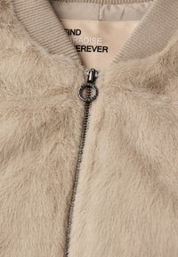 Beige fuskpäls bomberjacka med ribbade muddar och krage, med en silverfärgad dragkedja och tryckt etikett inuti. Mjuk textur och avslappnad design.