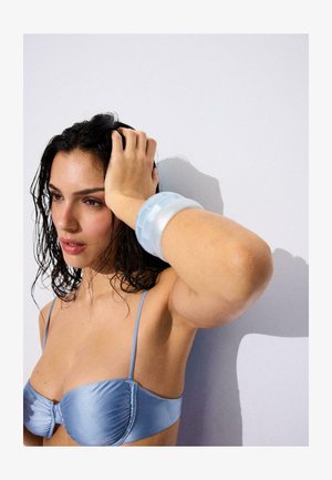 Žena s mokrom tamnom kosom u plavoj majici na tanke naramenice, nosi velike prozirne plave narukvice, odmara ruku na glavi na jednostavnoj pozadini.