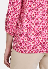 Kurzärmliges Top mit einem lebhaften pink- und cremfarbenen geometrischen Diamantmuster, ergänzt durch florale Akzente und einer lässigen Passform, kombiniert mit beigen Hosen.