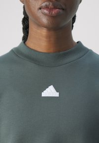 Olivegrön sweatshirt i bomull med ribbad rund halsringning. Har en vit Adidas-logotyp på bröstet. Slät textur.