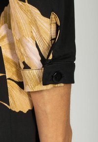 Mankiet czarnej koszuli z kwiatowym wzorem w beżowe i różowe liście, z czarnym guzikiem i kontrastującym ściegiem. Gładka tekstura materiału.