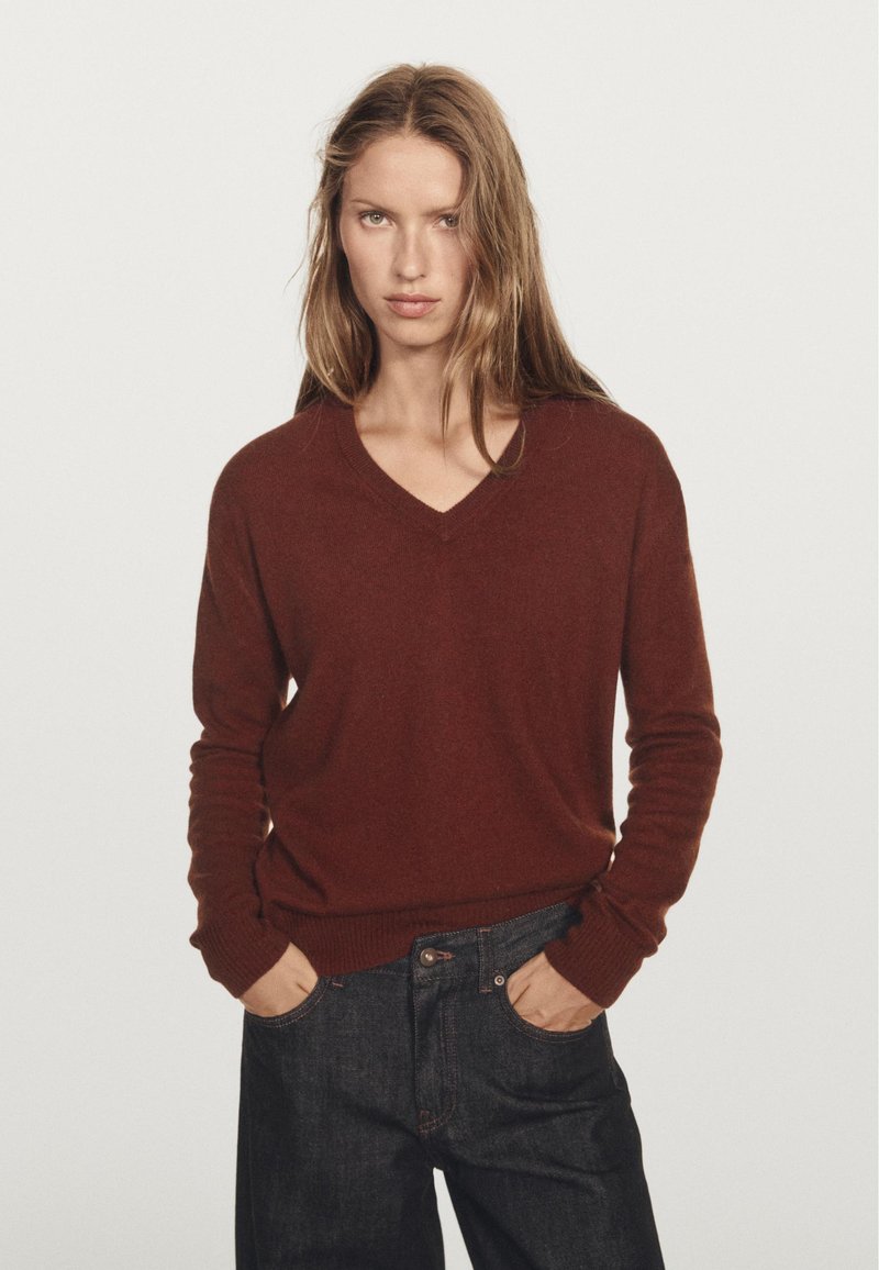 Pullover bordeaux a scollo a V realizzato in un materiale morbido, con maniche lunghe e polsini a coste. Indossato con jeans in denim scuro, mani nelle tasche.