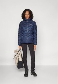 Moteris vilki tamsiai mėlyną pūstinį švarką, juodas kelnes ir žygio batus, laikydama šviesiai rudos ir juodos spalvos kuprinę už viršutinės rankenos.