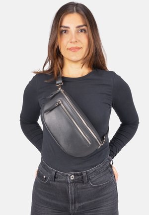 Frau trägt ein schwarzes Langarmshirt, schwarze Jeans und eine schwarze Leder-Umhängetasche mit zwei silbernen Reißverschlüssen quer über der Brust.