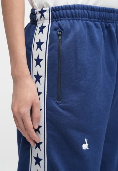 Pantaloni de trening bleumarin cu dungi laterale albe cu model de stele și buzunar cu fermoar. Prezintă un mic logo alb cu iepure în față, lângă buzunar.