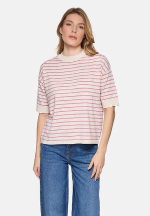 Mujer de pie contra un fondo liso, vistiendo un suéter crema de manga corta con rayas horizontales rosas y jeans azules.