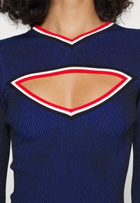 Top en tricot côtelé bleu avec un design en V et une découpe audacieuse, accentué par des bordures rouge, blanc et noir autour de l'encolure.