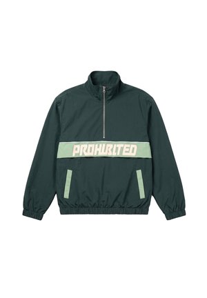 Mann trägt eine dunkelgrüne Halbreißverschlussjacke mit dem Text "PROHBITED" und eine schwarze Kappe mit weißer Schrift, steht vor einem einfarbigen hellen Hintergrund.