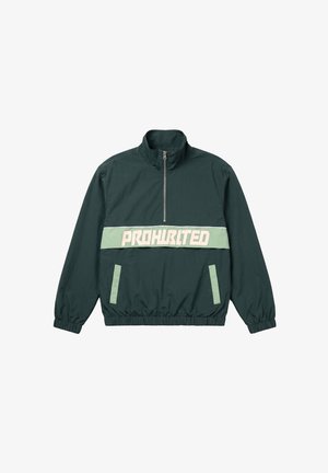 Mann trägt eine dunkelgrüne Halbreißverschlussjacke mit dem Text "PROHBITED" und eine schwarze Kappe mit weißer Schrift, steht vor einem einfarbigen hellen Hintergrund.