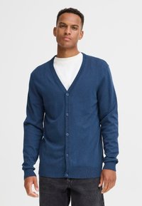 Cardigan da uomo blu navy, con scollo a V a coste, maniche lunghe, chiusura con bottoni, tessuto lavorato a maglia, abbinato a una t-shirt bianca a girocollo.