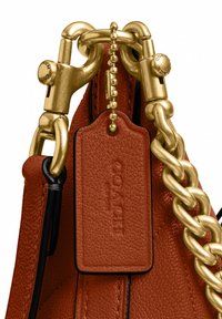 Gros plan d’un sac en cuir marron texturé avec de grands maillons de chaîne dorés et une étiquette en cuir assortie attachée par une chaîne à billes dorée.