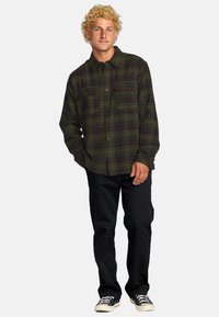 Camisa de franela a cuadros verde y negra con dos bolsillos en el pecho, con botones en la parte delantera, llevada con pantalones oscuros y zapatillas. Textura de tela suave.