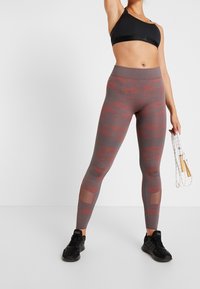 High-waisted maroon and gray camouflage leggings with mesh accents at the calves, paired with a black sports bra and black sneakers.