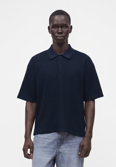 Hombre de piel oscura que lleva una camisa polo azul marino oscura de manga corta y holgada, y vaqueros azul claro, de pie frente a un fondo blanco liso.