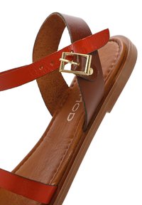 Sandalia de cuero marrón con correas rojas y un hebilla de metal dorado. Suela plana con una plantilla marrón suave. Diseño minimalista con punta abierta.