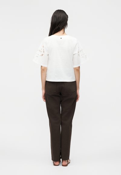 Γυναίκα με μακριά σκούρα μαλλιά, που φορά λευκό κοντομάνικο τοπ με κοψίματα και μαύρο παντελόνι ίσιας γραμμής, στέκεται γυρισμένη με την πλάτη.