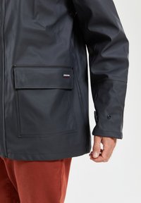 Zwarte waterdichte jas met een voorkantzak, drukknoopjes op de mouwen en een gladde rubberachtige textuur. Draagt over roestkleurige broek.