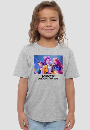 Smiling young girl wearing gray t-shirt featuring animated female K-pop characters with "KPOP DEMON HUNTERS" text below.