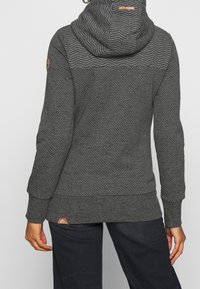 Persoon draagt een donkergrijze hoodie met een wit gestippeld patroon en een chevron-ontwerp op de bovenrug, gecombineerd met zwarte broek.