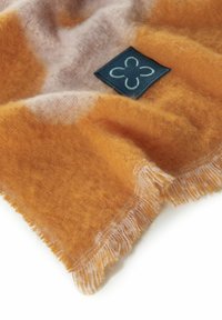 Oranje en beige geweven sjaal met een zachte textuur, voorzien van rafelige randen en een donkerblauw label met een bloemenontwerp.
