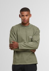 Jauns vīrietis valkā olīvzaļu krekli ar garām piedurknēm, uz kura ir teksts "SundayRun", stāv ar saliktām rokām pret vienkāršu gaišu fona.