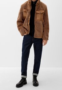 Mand iført brun fleecejakke med lynlås under en sort skjorte, mørke jeans med opkrav og sorte læder snørebåndsstøvler står op ad hvid baggrund.