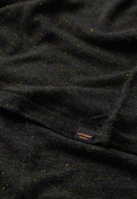 Zwarte stof met gele spikkels, zachte textuur. Bevat een klein "Superdry Premium" label dat aan de rand is gestikt.