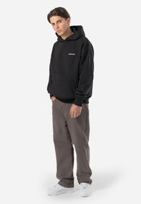 Svart hettegenser med frontlomme, med en hvit logo. Kombinert med løse grå bukser og hvite joggesko. Glatt tekstur.