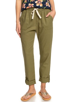 Person iført olivengrønne bukser med manchetter, forlommer og snøre i taljen, kombineret med metalliske thongsandaler og en blomsteragtig top.