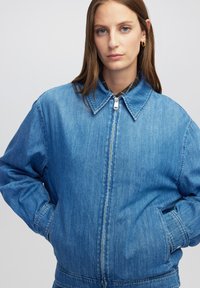 Jonge vrouw met lang bruin haar, draagt gouden oorbellen en een blauwe spijkerjack met rits, met handen in de zakken.