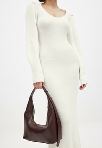Robe blanche en tricot à manches longues avec une texture côtelée et un col rond. Tient un sac à main en cuir marron lisse au design incurvé.