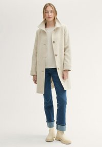 Beige gyapjú keverék kabát magas gallérral és három gombbal, csíkos felsőre, farmernadrágra és bézs bokacsizmára feltéve. Minimalista dizájn.